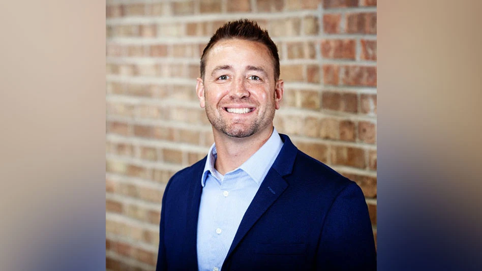 a smiling man with stubble on his face wearing a blue suit jacket and shirt standing in front of a brick wall